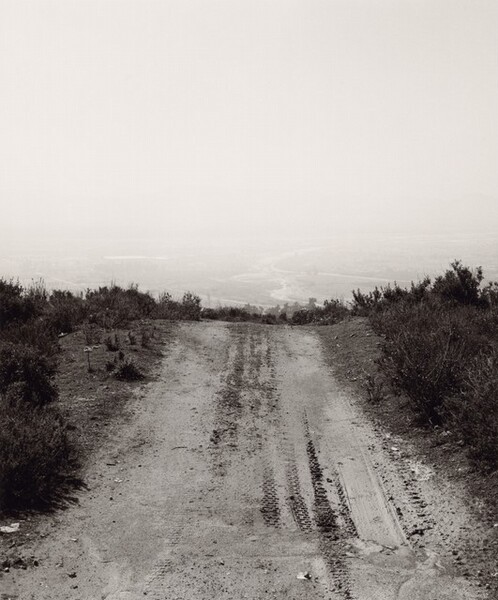 A mud road lined with tire tracks moves away from us up the center of this vertical black and white photograph. Scrubby bushes and vegetation line the road to each side. A band of treetops over the ridge gives way to a hazy landscape with a river winding through flat land.