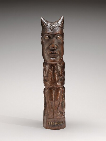 This stylized, free-standing, wood sculpture shows a seated man with horns, creating a column-like shape. The statue is carved from polished, reddish-brown wood. The column is divided into three sections for the head, torso, and legs. In this photograph, the man faces us. He has gold-colored eyes under arched brows, a wide nose, thin lips, and a square jaw. The horns rise from over his temples. In the middle section, he holds his hands in front of his chest, nearly touching as if in prayer. The bottom section shows his shins, knees together. His feet, which are roughly carved, turn outward. The base along the bottom is carved with words painted in gold, reading, “PERE PAILLARO.”