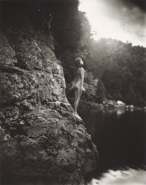 At the center of this composition, a naked girl stands poised at the base of a rocky cliff facing a body of water in this black and white vertical photograph. Light glints off her pale skin in a way that makes it look almost like burnished metal. Her dark hair falls over her shoulders in wet cords. Her body is angled away from us but we see her face in profile. She stands tall, her chin lifted, her eyes downcast, her lips together, perhaps blowing out a breath. Her arms reach straight by her sides next to her buttocks, and her hands are lifted outward, like small wings. She stands balanced on the rock’s edge, her feet together and her toes angled down. The body of water to our right is mostly ink black, but white clouds reflect in its shiny surface in the lower right corner. The girl and the rocky cliff behind her are in crisp focus. Blurry in the background, more tree-covered cliffs fill in most of the rest of the photograph, except for a patch of white sky in the upper right corner. Bright sunlight there washes out the trees nearby.