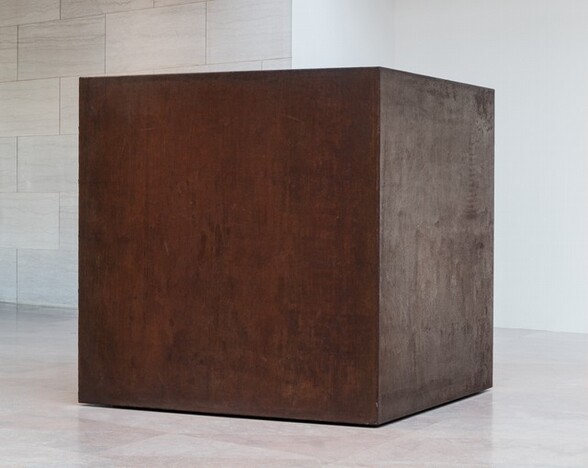 A rust-brown, steel cube sits directly on a pale pink stone floor in a gallery. This photograph shows the cube from near one of the corners so two sides are visible. The surface of the steel is mottled with bronze and and dark brown, and is faintly streaked. The bottom edge of the cube seems to float slightly, creating the impression that hovers just above the floor. The room around the cube has flat marble panels to the left and a white wall to the right.
