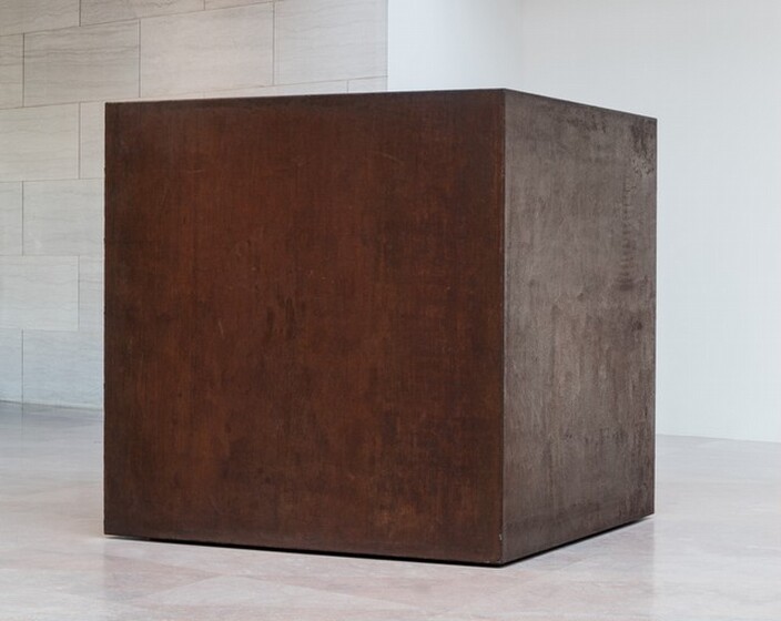 A rust-brown steel cube sits directly on a stone floor in a gallery. This photograph shows the cube from near one of the corners so two sides are visible. The surface of the steel is mottled and faintly streaked. The bottom edge of the cube seems to float slightly, creating the impression that hovers just above the floor. The room around the cube has flat marble panels to the left and a white wall to the right.