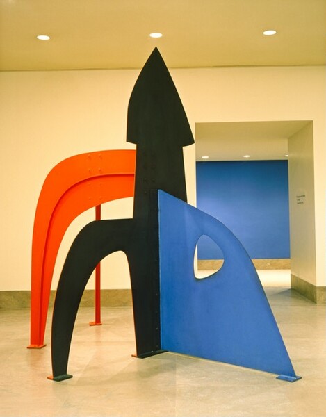 Three flat metal sheets cut into different shapes and each in a single color—black, red, or blue—make up this free-standing, abstract sculpture. This photograph shows the piece in a lobby space, and the top of the sculpture nearly touches the ceiling. At the center of the sculpture, a black, tall form like a backward-facing lowercase h is topped with a triangle reminiscent of an arrow. The short leg of the h faces our left in this photograph and is angled slightly toward us. To our left and affixed to the back side of the black arrow, a red form arches from near the arrowhead to the ground to create an empty space below. Seen only from the narrow edge in this photograph, another red piece arches back and away from us, down to the floor. Whereas one could conceivably walk or crawl under the black and red arches, the third blue piece to our right is a mostly solid sheet. It is attached to the center of the wide side of the arrow, facing us, and it moves toward us at an angle to our right. A rounded triangular form is cut out of the blue sheet near the upper right.