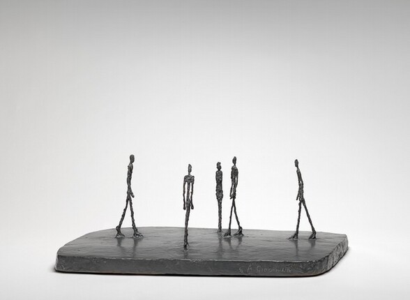 Four thin people with exaggerated, elongated legs, torsos, and arms walk across a flat, smooth, dark slab while one stands with feet together and arms by their side in this free-standing bronze sculpture. They are sparsely spaced across the base, and they all head for roughly the center of the slab. The person standing with feet together is at the center of this photograph, which shows the sculpture from one long end. The people’s bodies are textured and knobby, as if built up from small pieces of clay. The artist’s name is incised into the front face of the base to our right, and reads: “4/6 A. Giacometti.”