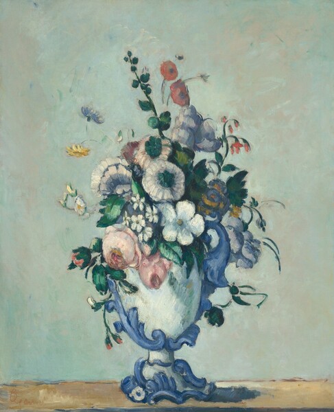 Rose and coral pink, vivid white, delphinium blue, and buttercup yellow flowers with deep green leaves are arranged in a blue and white vase in front of a pale sea-green background in this vertical still life painting. The vase has a white cup on a flaring foot. The vase is encircled with a twisting, decorative blue vine and scrolls. It sits on a band of peanut brown, suggesting a wooden tabletop. The still life is painted with blended strokes, giving the composition a soft, almost mottled appearance. The artist signed the painting in the lower left corner, “Cezanne.”