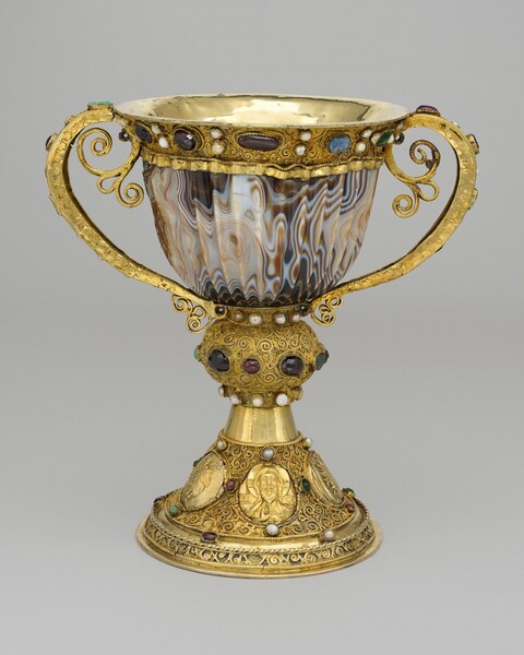 A cup carved from stone is mounted in gold fittings to create a jeweled chalice with handles that curl up each side and a tall, flaring foot below. The stone of the cup is swirled with shell pink and rust brown, and carved to create vertical ridges. The gold rim around the lip flares outward and is set with a band of alternating pairs of small, white pearls and mostly garnet-red stones, though one stone to our right is a muted blue. In this photograph, the handles curve up each side from the base of the stone cup to the gold lip, where the handle divides into two scrolling tendrils on either side of a teardrop shaped center, like curling petals. The base and foot are as tall as the stone cup and gold lip. A knob-like form just below the stone cup is set with deep red and jade green stones around its center, and rows of small pearls around the narrow top and bottom of the knob. The knob is chased to create a pattern of scrolling vines, like tracery. The flaring foot below is also chased with the same pattern, surrounding gold, oval medallions set at regular intervals in the foot. In our view, the medallion to the left shows a bunch of grapes and the center is a portrait of a bearded man with a halo, holding his right hand up with his first two fingers raised. The details on the medallion to our right are difficult to make out but it could be a sheaf of wheat. Small, round jewels are set above and below each medallion, and between each one.