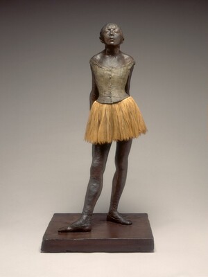 This sculpture is of a young girl in a ballet costume standing before us with her hands clasped behind her lower back, her head tipped back, and her chin thrust forward. The skin of her face, arms, chest, and legs are covered in bronze-brown wax, and her body faces us in this photograph. The sleeveless, putty-gray bodice of her costume fits tightly above a layered, fabric, pale, tea-brown tutu, and she wears ballet shoes. Her hair is pulled back but bangs sweep across her forehead, brushing her nearly closed, heavy eyelids. Her lips are slightly parted, and light catches the broad planes of her cheekbones. Her shoulders are drawn back by her pose, holding her hands behind her with arms straight, and her hips jut slightly forward. She stands with her right foot extended in front of her, with both feet turned out, parallel to each other. She stands on a square, wood platform.