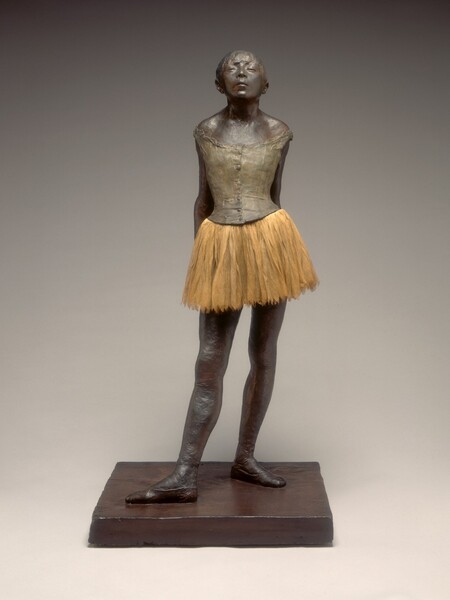 An adolescent ballerina stands before us with her hands clasped behind her lower back, her head tipped back, and her chin thrust forward. The skin of her face, arms, chest, and legs has been painted bronze-brown, and her body faces us in this photograph. The sleeveless putty-gray bodice of her costume fits tightly above a layered, fabric, pale, tea-brown tutu, and she wears ballet shoes. Her hair is pulled back but bangs sweep across her forehead, brushing her nearly closed, heavy eyelids. Her lips are slightly parted and light catches the broad planes of her cheekbones. Her shoulders are drawn back by her pose, holding her hands behind her with arms straight, and her hips jut slightly forward. She stands with her right foot extended in front of her, with both feet turned out, parallel to each other.