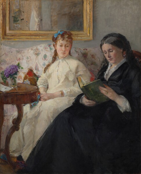 An older woman wearing a long black dress sits reading a book in front of a younger woman wearing cream white, who sits on a couch just beyond, in this vertical portrait painting. The scene is loosely painted with some visible brushstrokes, especially in the furniture, clothing, and room. Both women have pale skin and wear dresses with long sleeves and skirts. To our right, the older woman sits in a chair with a curved back, which runs parallel to the right edge of the canvas. The full skirt of her black dress angles down across the picture, almost spanning the bottom edge of the composition. Her gray hair is pulled back and covered by a black cloth or veil, which falls to her shoulder and blends with her dress. The collar is lined with a white ruffle, and white lace cuffs encircle her wrists. Her face is painted with rosy tones. She looks down at her book with dark eyes, and her rose-pink lips are closed. She wears a gold ring with a light stone on her left ring finger, and the book she holds has a moss-green cover. Her skirt overlaps the younger woman sitting nearby on the couch, which runs along the back wall of the room. The young woman’s knees are angled to our left but she looks slightly back to our right, to gaze down and into the distance with brown eyes. Her skin is paler, but she has lightly flushed cheeks and her petal-pink lips are also closed. An azure-blue bow is tied in her chestnut-brown hair. A few tendrils fall down her back and over one shoulder. Her dress falls loosely over her torso and legs. She wears a gold ring on her left hand and a gold bracelet on that wrist. She fingers a piece of lapis-blue fabric in one hand. The couch behind her has a pattern with stylized flowers or leaves floating within rose-pink ribbons that hang in swags, against a white background. Pillows patterned with ruby red, sky blue, and fawn brown peek out from behind the young woman. A round or oval wooden table with curving legs to our left holds a glass vase with a violet-purple flower, perhaps a hydrangea. A slip of paper or envelope sits on the table next to the vase. The wall behind the couch is fog gray, and the bottom edge of a gold frame hangs behind the young woman.