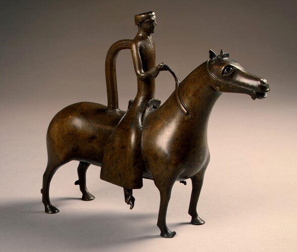 This free-standing bronze sculpture shows the rounded, stylized forms of a person with long limbs riding astride a horse with noticeably short legs. The horse and rider are angled to our right in this photograph. The dark brown bronze gleams white where the light catches the smooth surfaces, and some areas are a burnished gold color. The horse looks ahead with large eyes. It has short ears, and its mouth is parted. The person riding the horse holds the reins with the right hand, closer to us. The reins wrap halfway around the horse’s neck and are then broken off. The cleanshaven rider has a heavy brow ridge and a long, straight nose. He wears a flat-topped cap and a robe that reaches to his ankles. The pointed toes of his shoe pokes through the small stirrup we can see. A rounded handle connects the rider’s upper back to the back of the horse. The background is taupe-gray behind the sculpture and darkens at the upper corners.