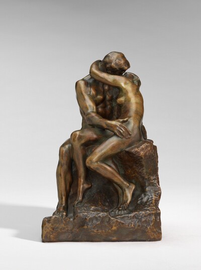 A man and woman embrace and kiss while sitting on a rocky formation in this free-standing bronze sculpture. Both people are nude, and they turn toward each other. In this photograph, to our right, the slender woman reaches her left arm, closer to us, up to wrap around the man’s neck. Her left breast is silhouetted against the man’s muscular chest. The woman’s raised arm covers most of their faces but the man turns his head down to meet hers. The man rests his right hand, closer to us, on her hip and she hooks one leg over his. His knees are angled to our left and he turns his torso toward her. The woman’s left toes brush his foot, just below hers. They sit on a textured, rock-like form. The surface of sculpture has a golden-brown patina, which darkens where the bodies fold and crease. The artist’s name is stamped into the seat, just below the woman’s hip: “RODIN.”