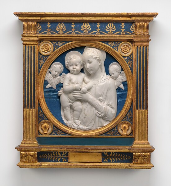 Sculpted in painted terracotta, a woman is shown from the waist up, bracing a standing nude, plump baby boy. The woman, baby, and two winged heads floating to either side are sculpted in snow-white relief against a flat, cobalt-blue background, all contained within a round gold frame. The roundel is then framed within a rectangular wooden panel ornamented with gold and blue molding, rosettes, and leafy designs. The boy, woman, and winged heads all have plate-like halos, and their eyes have hazel-brown irises outlined in black. Within the round frame, the boy stands to our left of center, and his halo nearly reaches the top of the roundel. His body faces us as he stands on his right leg, to our left. That foot seems to rest on the ledge of the round frame, and he leans against the woman, to our right. He looks off over our left shoulder, his pursed lips set between chubby cheeks. With both pudgy hands, he holds onto the woman’s thumb or garment as she braces his torso. Her body is angled toward the boy, and she looks down and off in that direction. She has a straight nose, a pointed chin, delicate lips, and her thin eyebrows are black. Her wavy hair is parted down the middle and pulled back under a veil, and her long-sleeved garment falls in folds down her front. Her other hand wraps around the baby’s far hip. The winged baby heads hover at her shoulders, one to each side. The blue background has some bands of lighter and darker blue, suggesting a cloud-streaked sky. Gold rosettes fill the top corners between the round frame and the square panel into which it is set. Shield-like forms fill the lower corners. Fluted columns rise to either side of the central panel, and molding runs across the top and bottom. Some of the molding is painted gold, while other areas have gold, leafy patterns painted against the blue background.