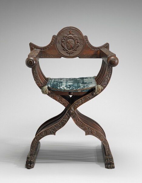 The curving X-frame design of this wood chair creates an hourglass silhouette from the front, and creates a bucket-like seat for the sitter. The front face of the wooden frame is carved with curving vines and stylized bunches of grapes and leaves, and the frame ends with clawed lions’ feet at the bottom. The seat and legs are made up of alternating slats so the chair could fold up. Imagine interlacing your fingers and then moving your palms back and forth—that is how the pivot of this chair works. The arms of the chair, which have a rounded knob at each front end, rest along the top edges of the slats. The back rail connects at the back of the arms. A crest at the center of the back rail is flanked by the capital letters “A” to the left and “G” to the right. The crest and letters are contained within a medallion-like form, and the sides of the back rail dip down to meet the arms. A worn, pale blue velvet cushion rests on the seat, and has tassels at the front corners. The chair is shown against a pale gray background.