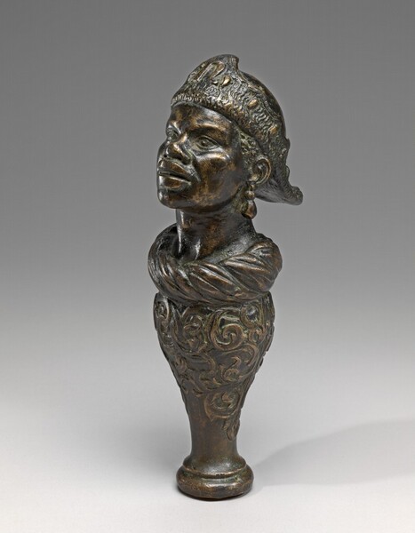 This free-standing, bronze sculpture shows the head and neck of a person atop a spindle-shaped base. The face is angled to our left in this photograph. The bronze surface has aged to an almost gold-colored patina, especially along the ridges and rounded surfaces. The person’s upturned face has large, almond-shaped eyes, full lips, high cheekbones, and a wide, upturned nose. The person wears a hat with a wide band flat across the forehead, and it flares like a duck bill at the back. Twisted fabric makes a ring that encircles the base of the neck and a large, teardrop shaped pendant earring hangs from the left ear, closer to us. The baluster-shaped pedestal is decorated with swirling scrolls.