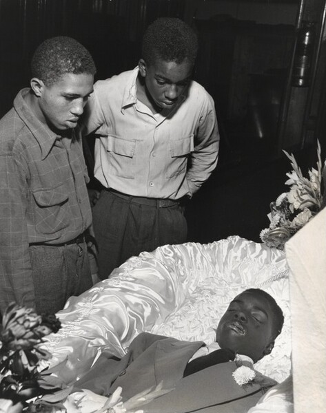 Two young Black men look at a third, who lies in a coffin, in this vertical black and white photograph. All three are clean-shaven with short hair. The coffin is lined with light-colored satin. We see the man there, Maurice Gaines, from the chest up. He wears a suit jacket, button-up shirt, and dark tie. A flower is pinned to the lapel of the jacket. His skin is dark, and there are some lighter spots around his mouth. Flowers are next to and on the edges of the coffin at his waist and at the head. The two onlookers both wear button-down shirts with pockets with flaps on the chest. They look steadily at Maurice, mouths parted. One leans forward with his arms planted on the back of his hips. The other looks down with the arm we see hanging by his side. The three men take up most of the composition, and the background behind them is inky black.