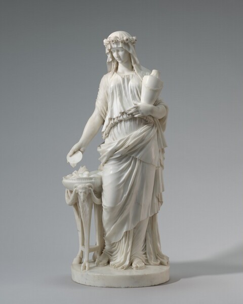 Carved from cream-white marble, a woman stands next to a hip-high, three-legged table in this free-standing sculpture. Her weight rests on her left leg, her right knee bent. She wears a flower crown over a garment that covers her head and falls in sweeping, voluminous folds to her sandaled feet. An urn is tucked into her left elbow and she holds a saucer-like dish in her right hand down by her side to pour oil onto the flames on the altar below. She looks down toward the altar, which is decorated with a ram’s head and garlands.