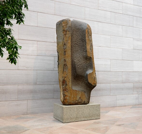 A rough-hewn block of basalt sits upright on a rectangular stone base on a pale pink stone floor against a stone wall. In this photograph, we are off the front left corner of the free-standing sculpture in a space filled with natural light. The tongue-shaped basalt is narrower from front to back and wider from side to side. It is about three times taller than it is wide at its widest. Some of the dark brown basalt running up the center of the block is smooth but most is pitted. Strips up either side have been carved to expose rough, copper-colored stone, some of which has flaked away. The tan stone base is speckled densely with gray, and the leaves of an indoor but mature tree bow into the top left corner of the photograph.