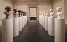 Creating two lines that move away from us, a row of seven busts of a woman’s head and neck made from brown chocolate face a row seven busts showing the same woman made from white soap, each on an identical white, columnar pedestal. In this photograph, they are placed in a long room with cream-white walls, tan stone molding, and a dark gray marble floor leading to an open doorway at the far end of the room, across from us. Each bust shows a woman with a small button nose, pursed lips, and closed eyes. Her hair is pulled back in a bun at the nape of her neck. Each bust ends just below the shoulder line and is held on a base that flares out like a chess piece to act as a foot. Each sits on a columnar pedestal that comes about a third of the way up the height of the tall doorway. Though the faces look similar or identical at first glance, closer inspection shows that some are worn at different areas, like the chin, forehead, nose, cheeks, or bun. One of the soap busts, at the far end, is missing the entire crown of the head and the profiles of two more soap busts and one chocolate bust are worn down so much it appears the face is missing. The surface of some of the chocolate busts looks almost frosted where the light hits it. The ivory color of the soap busts are more consistent.