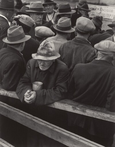 More than a dozen men crowded together fill this vertical black and white photograph. All wear snap-brim hats, fedoras, or caps, and all but two away from us. One man in the crowd looks to our right. The other is closest to us. He rests his forearms on a wooden railing, a metal cup tucked between his elbows. His hands are tightly clasped, and his worn, stained fedora is low over his lined face. His thin lips are set and he has a grizzled white beard. A partially legible sign in the back reads 