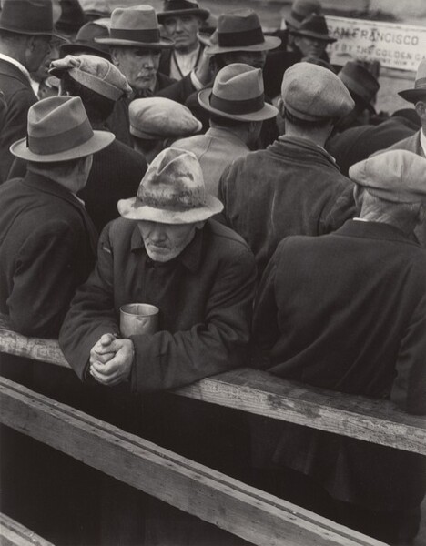 More than a dozen men crowded together fill this vertical black and white photograph. All wear snap-brim hats, fedoras, or caps, and all but two away from us. One man in the crowd looks to our right. The other is closest to us. He rests his forearms on a wooden railing, a metal cup tucked between his elbows. His hands are tightly clasped, and his worn, stained fedora is low over his lined face. His thin lips are set and he has a grizzled white beard. A partially legible sign in the back reads SAN FRANCISCO and BY THE GOLDEN GA.
