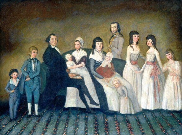 Twelve men, women, children, and babies are arranged on or around a couch in this horizontal portrait painting. The features and clothing are simply painted, so some of the faces are a bit generic. They all have pale skin with rosy cheeks, and all but one of them have blue eyes. The slate-blue couch is centered against a wall mottled with copper brown, golden yellow, and sage green. Three adults sit on the couch. To our left, a man wearing a black suit and stockings leans against the arm of the couch so his body faces our right in profile, his legs crossed. He turns to look at us with a slight smile on his lips, his eyebrows raised. His dark hair is smoothed back along the top of his head and is bushy over each ear. A white neckerchief wraps across his high collar and  ruffles cascade down the front. He holds a book in his lap. Two women sit next to him, to our right. Both have dark hair and wear broad neckerchiefs over tightly fitting bodices, and white aprons over long, full skirts. The woman to our left turns her face slightly to our left but looks at us from the corners of her eyes. She wears a white bonnet, and her bodice is plum purple. The other woman’s head is uncovered and her forehead lined with bangs. Both of these women hold babies wearing white caps, and the babies smile. Just behind the couch, to our right of the second woman, a young man with a faint, dark beard, black hair, and brown eyes stands with his body facing our left in profile. He turns to look at us as he tucks one hand between the buttons of his nickel-gray, high-collared jacket. Two boys wearing blue suits stand to our left in front of the couch, near the seated man. The smaller boy, to our left, holds an archery bow, wears a dark cap, and looks at us, smiling. The older boy standing next to him has blond hair and points down at the younger child with one hand, the other on his hip. Two young women and a girl stand to our right, also in front of the couch. They all wear white dresses with long sleeves and skirts, their narrow waists tied with rose-pink sashes. Two older girls have black hair and hold hands. The youngest girl, near the right edge of the painting, has blond hair and also looks at us. The carpet beneath the group has vertical stripes leading back to the wall. Narrow bands of navy blue are dotted with pink to suggest a floral pattern, between wider, light blue stripes.