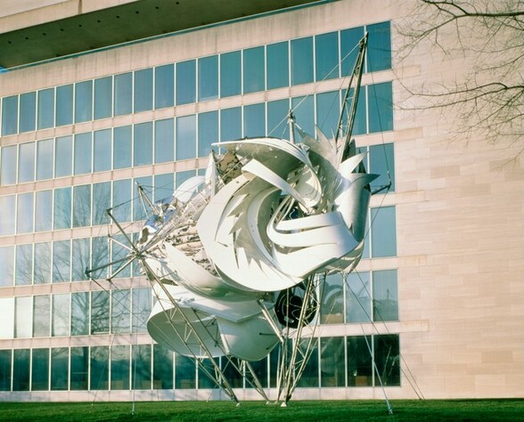 This freestanding, mostly white and silver metal sculpture stands as tall as five of the six stories of windows in the building behind it in this photograph. The sculpture is made up of swirling white forms, like cut and curled paper, wrapped around and among silver rods, struts, and rigging-like cables. Four silver legs gather in a narrow spot on the grassy ground and angle up and outward around the collection of central, white forms. The white sheets are cut into zigzagging lines and straight slits, and then curl around themselves. Other material is nestled in among the white sheets and metal bars. This includes a material mottled with pale pink and dark gray, built up like clay rolled into coils and stacked near the center. A star-like, silver form flares out at the upper right. Taut cables anchor the piece to the ground and connect the legs of the framework. The piece sits on green grass before a pale peach stone building with reflective blue-gray windows.