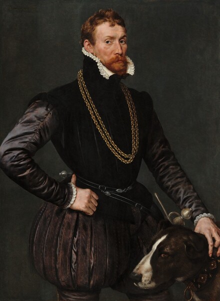 Shown from the thighs up against a dark background, a man with light skin, wearing a black satin jerkin with puffed breeches, stands with his hand on the neck of a dog by his side in this vertical portrait painting. The man stands with his body angled to our right but looks at us from the corners of his dark eyes. His brows are subtly raised, and his cheeks are slightly sunken under high cheekbones. His red hair is cut short, and his red mustache and beard are trimmed. The high collar of his black jacket is lined with a white ruffle along his chin line. Light from our left glints off the long sleeves of his tight-fitting jacket, creating the impression that it is made of heavy silk. Two thick gold chains hang down his chest, and a sword hangs from a belt around his waist, the hilt on the hip farther from us. His pants puff out and gather on the mid-thigh over stockings. He hooks the thumb of his right hand, on our left, around his hip and he rests his other hand on the neck of the dog standing by his left side, to our right. The dark brown dog has a white stripe down its nose. It faces our left in profile but its brown eyes look toward us. On the collar, ornate metal bosses alternate with low spikes, set in a wide black band. A ring hangs from the front of the collar. Barely legible, the painting is signed and dated in the upper left corner with dark paint: “Antonius mor pingebat a. 1569.”
