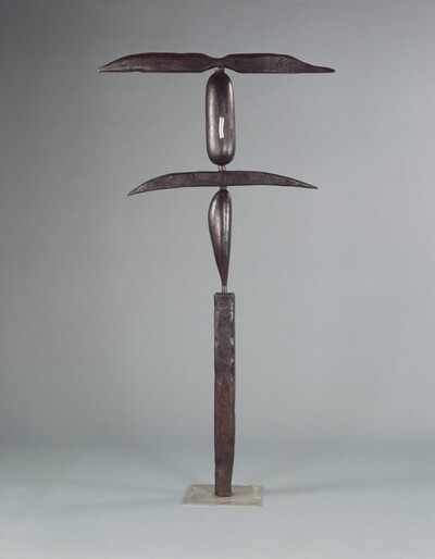 This tall, abstract, bronze, free-standing sculpture is made up of five thin, elongated geometric shapes stacked vertically. The surface of the bronze is dark, pewter gray, and it is photographed against a lighter gray background. The sculpture stands on a square, plate-like, silver-colored base. Starting at the bottom, the first section is a tall, narrow, rectangular column with a pitted surface and irregular edges, as if the corners had been hammered. The next vertical section is a slender, smooth, leaf-shaped object that swells at the top and tapers to a point where it meets the column below. A wide, narrow sliver balances horizontally across the leaf-like object, and it tapers to points to our left and right. Above, a vertical capsule-shaped form is painted with two small, vertical, wavy lines in white. Finally, another horizontal form with a hammered surface tops the structure. It is pinched at the center and swells slightly to either side before tapering to points. The leaf form and capsule shapes are about the same height. The topmost horizontal piece is slightly wider than the lower one.