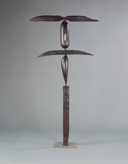 This tall, abstract, bronze, free-standing sculpture is made up of five thin, elongated geometric shapes stacked vertically. The surface of the bronze is dark, pewter gray, and it is photographed against a lighter gray background. The sculpture stands on a square, plate-like, silver-colored base. Starting at the bottom, the first section is a tall, narrow, rectangular column with a pitted surface and irregular edges, as if the corners had been hammered. The next vertical section is a slender, smooth, leaf-shaped object that swells at the top and tapers to a point where it meets the column below. A wide, narrow sliver balances horizontally across the leaf-like object, and it tapers to points to our left and right. Above, a vertical capsule-shaped form is painted with two small, vertical, wavy lines in white. Finally, another horizontal form with a hammered surface tops the structure. It is pinched at the center and swells slightly to either side before tapering to points. The leaf form and capsule shapes are about the same height. The topmost horizontal piece is slightly wider than the lower one.