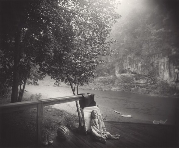 In the lower center of this black and white, horizontal photograph, a light-skinned child sleeps sitting up against a folded-up lawn chair by the banks of a river. Small in scale against the landscape, the child’s long hair is wet and the eyes are closed, mouth hanging slightly open. A patterned towel is wrapped around the child’s shoulders and meets below the waist, showing a V of bare chest and belly. The towel drapes over the legs. The child sits near the edge of a wood planked deck or dock. The collapsed folding chair has three parts, and it pins a shiny black object, presumably a deflated inner tube, against the post of the railing. The railing extends across the left half of the composition and off that edge. A pair of sneakers and a watermelon sit next to a post supporting the railing halfway along its length. Beyond the deck to our left, the dirt riverbank has some patches of grass. The round canopy of a tree there fills the top left quadrant of the photograph. The river angles toward us from the back left, and the far bank is lined with scrubby brush and trees growing up and over steep, rocky outcroppings. An inflatable alligator and raft sit at the water’s edge on the bank closer to us, to our right of the sleeping child. An unfolded deck chair sits beyond the deck, farther along to our right. Bright sunlight blurs the view at the top center of the photograph and the scene gets darker at the edges and corners. The photograph goes in and out of focus, so the back of the folding chair behind the child and some of the leaves on the tree are in the sharpest focus. The child is slightly blurry as are the other objects and toys. The image becomes notably blurry at the edges.