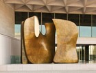 A gleaming, abstract bronze sculpture made of at least two rounded forms fills a two-story, covered entryway. The rounded, smooth forms sit side-by-side. The form on our right curves slightly inward up the left side and is lightly pinched at the center along the right side. The form on the left is shaped like a fat U with a deep, wide curve at the bottom. Most of the sculpture’s surface is lightly mottled in a rich honey color, except at the top of the front face of the U’s arm, which looks as if it were cut vertically to expose an elongated oval shaped disk of highly polished, bright gold-colored bronze. A third form appears as a straight vertical element behind and to the left of the U-shaped piece. The building behind and over the sculpture has two rows of floor-to-ceiling windows, which reflect darkly in this photograph. The floor and ceiling above are smooth, buff-colored stone. Two rows of deep-set triangles recede into the ceiling.