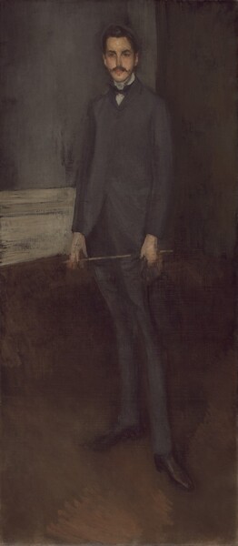 A tall, thin, pale-skinned young man stands in a dimly lit room, nearly filling the height of this portrait painting. He faces us with his body turned slightly to our left, and he wears a charcoal-gray suit and black shoes. A black bow tie and silver-gray shirt collar are visible above his buttoned jacket, and he holds what might be a riding crop in his elegant, long fingers. His short hair, moustache, eyebrows, and eyes are all chestnut brown. His hair is swept off his forehead and cut around the ears. Thick eyebrows frame his large eyes, and a handlebar moustache curves over full, peach-colored lips. He stands near the corner of a room. The brown floor seems tipped toward us, so the parchment-white baseboard runs behind the man at hip level. The wall to our left is stone gray, streaked with darker gray. The right wall blends with the dark brown of the floor, which is loosely painted with patches of sable brown. Much of the portrait is loosely painted, especially in the man’s hands, clothing, and the room around him.