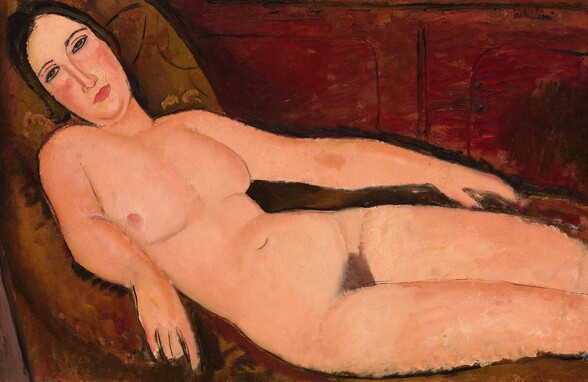 A nude woman lying across a brown divan in front of a russet-red wall is loosely painted with areas of mottled color in this horizontal composition. The woman fills the wide canvas so her head is angled into the upper left corner, and her legs extend off the right edge of the composition below the knees. Her long, oval face rests back against the couch, and she looks at us with slitted, almond-shaped eyes under dark, shallowly arched brows. She has a long, straight nose, flushed cheeks, a double chin, and her pink lips are closed. Her breasts fall to each side so we only see the pink nipple closer to us. She has a rounded belly and a triangle of brown hair at her groin. Her arms rest alongside her body. The couch has a low, upholstered arms sweeping down the sides. Black lines in the red wall behind the couch suggest molding or cabinetry. The artist signed the painting in the upper right corner, “modigliani.”