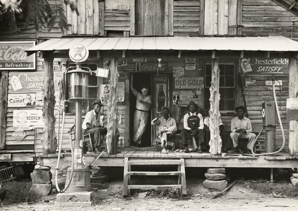 <p>Dorothea Lange, Country store on dirt road. Sunday afternoon. Note the kerosene pump on the right and the gasoline pump on the left. Rough, unfinished timber posts have been used as supports for porch roof. Negro men are sitting on the porch. Brother of store owner stands in doorway, Gordonton, North Carolina, July 1939, printed later