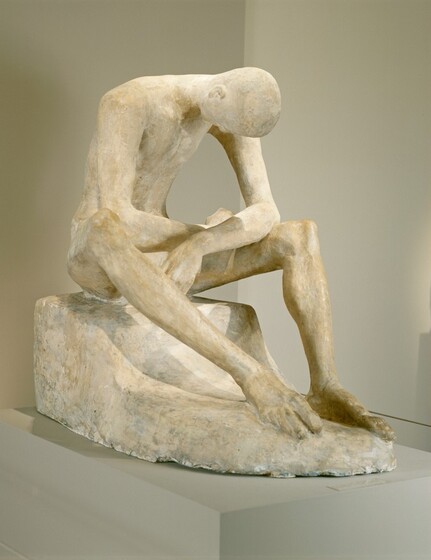 A man bows his bald head deeply as he rests his splayed elbows on his thighs in this freestanding plaster sculpture. The long toes of the man’s feet nearly touch, and they are angled to our right in this photograph. He sits on a low surface, also sculpted from plaster, and his long limbs create a cage around his hollowed chest. The surface of the plaster is ivory white in some areas and parchment brown in others. The piece rests on a gray pedestal against gray walls.