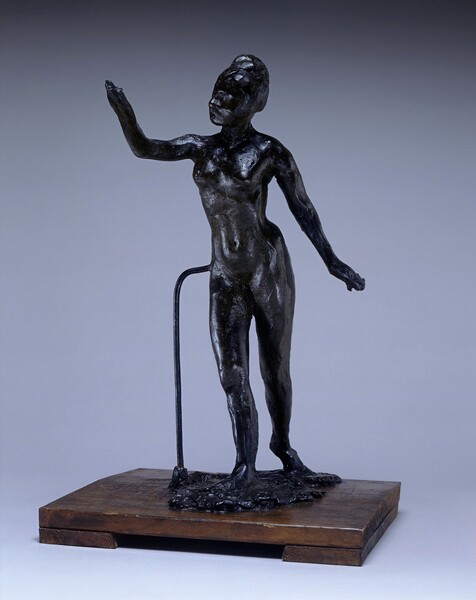 This standing nude woman is sculpted from black-tinted beeswax on a wood plank base. Her body is supported by a metal rod that connects the base to her right hip, to our left. Her hair seems to be pulled up and her facial features are indistinct. She steps forward onto her right foot, on our left in this photograph, so that the toes of her other foot touch the pool of wax on the base behind her. She lifts her right arm, on our left, in front of her so her upper arm is parallel to the base, and seems to look toward that hand. Her other arm is held away and slightly behind her body. Light reflects off some areas of the wax, which are smooth, while other areas are rough. The background lightens from charcoal gray along the top to nickel gray along the bottom of the photograph.