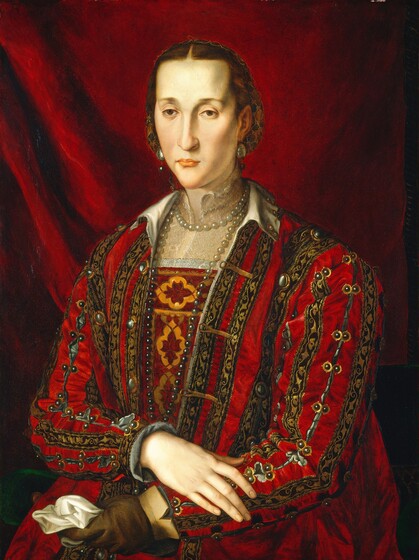 Shown from the hips up, a pale-skinned woman with a long, narrow face wears a richly decorated crimson-red dress in this vertical portrait painting. Her body angles to our left, and she looks in that direction with hooded brown eyes. She has a long nose, a pointed chin, and her pink lips are closed. Deep indentations at her temples and the hollows of her cheeks are shadowed. Her brown hair is parted down the middle and held back in a gold net. Large pearls hang from her ears, and a long strand of pearls doubled around her neck look white at her collar and gray down by her waist. The bodice of her dress is patterned with marigold orange and wine red, and translucent white lace covers her chest and flares up the sides of her neck. A heavy outer garment has black stripes decorated with gold leafy designs alternating with ruby-red stripes. The seams are studded with pearls down the front and along the split sleeves. A gray cuff lines the wrist of a bare, long-fingered hand, which rests across the other forearm. She wears a leather-brown glove on the other hand and clutches a white cloth in that fist. She sits on a pine-green cushion, and an apple-red curtain falls in gentle folds across the background.