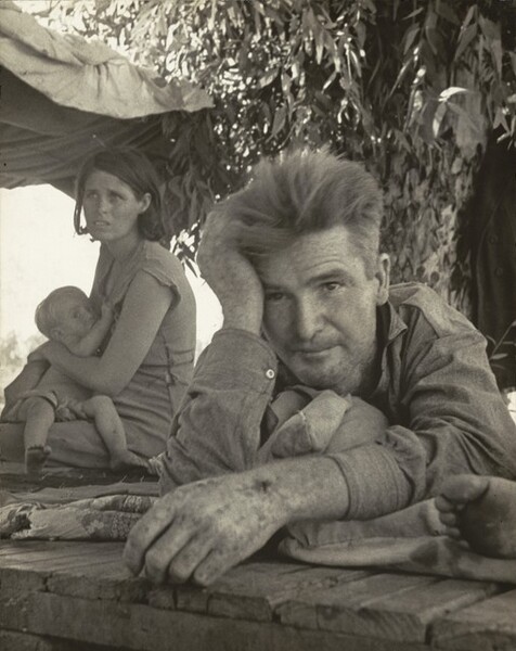 In this black and white, vertical photograph, a man, woman, and baby, all with pale skin, rest on blankets, or perhaps rugs, spread over a roughly built wooden platform under a leafy tree. Closest to us, the man takes up most of the right half of the composition. He lies on his stomach with his left arm, on our right, stretched out in front. His hand is close to the photographer so is slightly out of focus, and he cups his forehead with his other hand. The wooden platform on which he lies runs close to the bottom edge of the image. His light hair stands straight up from his head, and he is cleanshaven. He wears a long-sleeved, button-down shirt and looks off to our left and a little above us from under raised eyebrows and a creased brow. To our left, a woman sits beyond the man with her body angled to our left. Her chin-length dark hair is tucked behind her ears, and her worn dress has tattered seams and gapes at the waist. She hunches slightly over the child she holds and looks up and to our left, her mouth open and her eyebrows knit. She seems to breastfeed the young, barefoot child who clutches her dress. The baby looks tpward us from the corners of wide eyes. The toes and the ball of a person’s foot, mostly cropped from the image, are next to the man, along the right edge of the photograph. A piece of cloth is tied into the branches above and over the woman to create a makeshift tent against the bright glare of the sun.