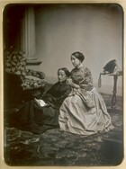 Two light-skinned women wearing full, floor-length dresses sit angled to our left at the center of this vertical black and white photograph. The woman to our left sits on a patterned carpet wearing a black dress and holding an unfolded piece of paper in her lap. The woman to our right is wrapped in a flowery shawl worn over a lighter colored dress, and she leans against the shoulder of the woman to our left. Both look into the distance to our left. There is a chair upholstered possibly with a floral-patterned fabric in the background to our left and a side table holding a bonnet, the ribbons falling over the edge, to our right. 