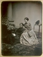 Two pale-skinned women wearing full, floor-length dresses sit angled to our left at the center of this vertical black and white photograph. The woman to our left sits on a patterned carpet wearing a black dress and holding an unfolded piece of paper in her lap. The woman to our right is wrapped in a patterned shawl worn over a lighter colored dress, and she leans against the shoulder of the woman to our left. Both look into the distance to our left. There is a chair upholstered with a floral-patterned fabric in the background to our left and a side table holding a bonnet, the ribbons falling over the edge, to our right. The women are mostly in focus and the rest of the room is a bit blurry, out of focus.