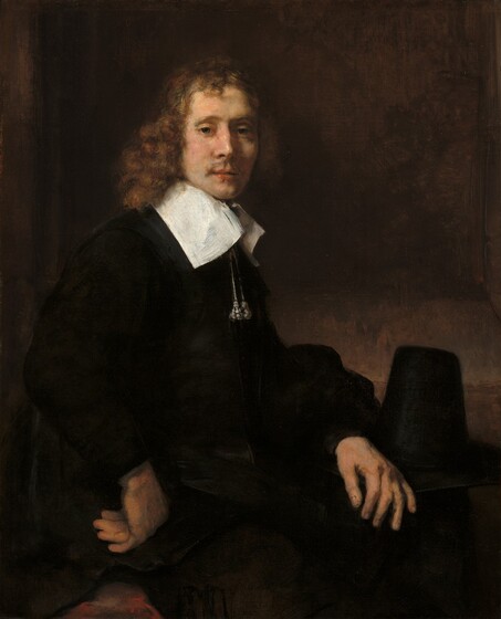 Shown from the hips up, a young man with pale skin and wearing black clothing sits with his body angled to our right as he turns to look at us in this vertical portrait painting. He looks out with green eyes under hooded lids. Light falls across his face from the upper left so his bumped nose casts a shadow on his high cheekbone. He has a wispy mustache over full, pink lips, which are slightly parted. His hair falls to his shoulders in auburn-brown curls, framing his slender face. Two silvery tassels hang on cords from a tall, wide, flat, bright white collar, which contrasts with the velvety black of his voluminous, long-sleeved jacket. His left hand, farther from us, rests on his knee and the other hand is curled so the back of his knuckles press against his right hip, closer to us. A patch of crimson red near that hand could be a pillow on his chair. His hat rests, presumably on a table, just beyond his legs. The hat has a tall, tapering, conical crown with a flat top. The shadowy wall behind him is mottled with slate gray and caramel brown, with a slightly darker panel to suggest a shallow niche.