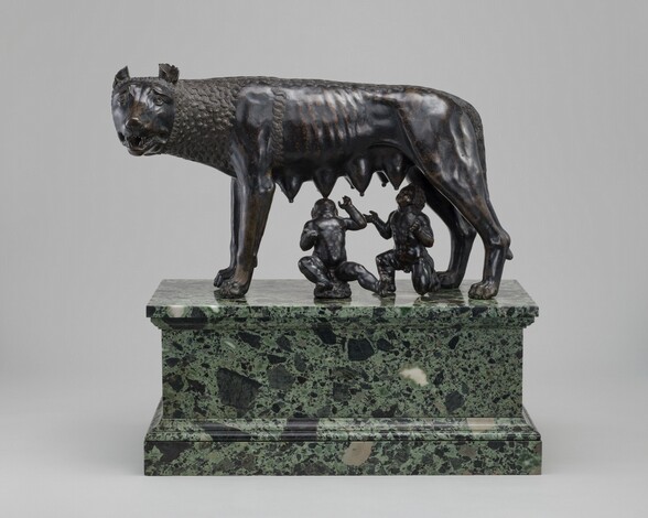 A free-standing, bronze she-wolf stands as two male infants attempt to nurse at her teats in this sculpture. In this photograph, the wolf stands sideways facing our left with her head turned to look at us, six teats hanging heavily along her underside. Her wide eyes are framed by raised but furrowed brows, and cup-like ears curl back on her round head. Her jaws are slightly open revealing sharp teeth. Her lean body is partly covered by tight clusters of parallel, undulating lines around her head, neck, and along her back, suggesting fur. Beneath her, one boy sits, rocking back on a low rock, and the other kneels. Both tilt their heads back and spread their arms wide as they reach for her teats. The bronze of the boys and wolf are rubbed gold in some areas, especially on the wolf’s ribs. The trio rest on a green and black marble pedestal.