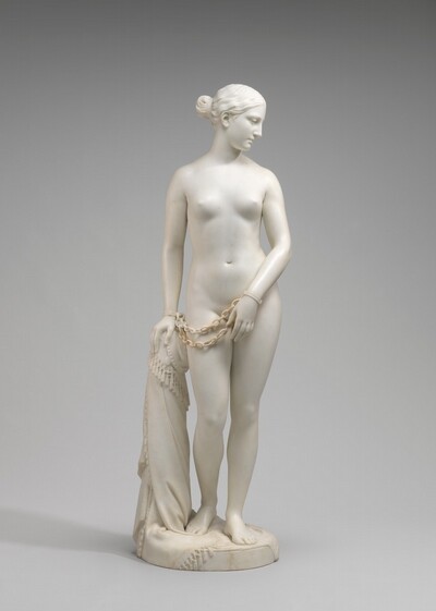 Carved from creamy white marble, a nude woman stands next to a hip-high support, perhaps a low post. In this photograph, her body faces us, and she looks down to our right in profile. Her wavy hair is tucked behind her ear and drawn back in a bun at the nape of her neck. Her weight rests on her left leg, on our right, and her other knee is bent. Her left arm is angled in front of her body so her hand covers her groin. Her other hand, on our left, rests on the post. Chains hang from shackles encircling her wrists. The post is covered with a cloth that gathers around the top and spirals to the ground beneath her feet, the edge trimmed with tassels. A cross and medallion peek out from under the cloth near her hand. She stands on a circular base.
