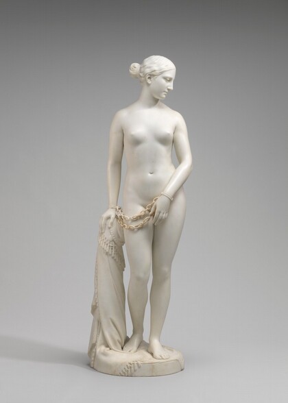 Carved from creamy white marble, a nude woman stands next to a hip-high support, perhaps a low post. Her weight rests on her left leg, on our right, and her other knee is bent. Her left arm, on our right, is angled in front of her body so her hand covers her groin. Her other hand, on our left, rests on the post. Chains hang from shackles encircling her wrists. She looks down to our right and is seen in profile in this photograph. Her wavy hair is tucked behind her ear and drawn back in a bun at the nape of her neck. The post is covered with a cloth that gathers around the top and spirals to the ground beneath her feet, the edge trimmed with tassels. A cross and medallion peek out from under the cloth near her hand. She stands on a circular base.