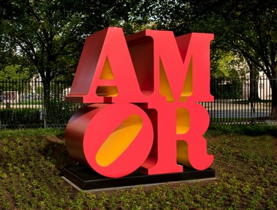 Four deep, three-dimensional letters spell out the word AMOR in this free-standing sculpture, with the A and M stacked on top of the O and R to create a square on a low black platform. The letters are coral red with butter-yellow undersides. The elongated, oval-shaped opening within the circular letter O is angled 45 degrees, toward the M at the upper right. We stand slightly to the left in this photograph so we can see the deep sides of the letters. The sculpture is displayed out-of-doors with trees and a tall black fence in the background and plantings around the base.