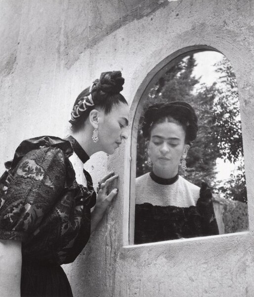 In this black and white photograph, a woman wearing a black dress leans toward an arched mirror set into a wall, so we see the woman twice in this double portrait. We are situated slightly below her so we look up at the woman, who gazes down and to our right. The surface of the wall is smooth but lightly textured, like adobe. Trees reflected in the mirror indicate that this is outdoors. The woman in front of us takes up the left half of the composition. She faces our right in profile and leans to our right, at the far edge of the mirror. Her dark hair is pulled up in ribbon entwined braids. She wears long, chandelier style earrings and her high-necked black dress has puffed, lace sleeves. We see her face straight on in the mirror’s reflection. The woman’s distinctive brows knit together over her rounded nose.