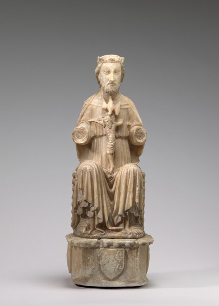 A seated man wearing a long robe and a crown has a crucifix in his lap in this free-standing, cream-white, alabaster sculpture. In this view, the seated man’s body faces us, and his arms are broken off beyond the elbow. He has short, wavy hair and a trimmed beard. His almond-shaped eyes are blank, and his lips are closed. His robe is deeply pleated and hangs in a U shape between his knees. A bird in flight, facing downward, hovers at his throat over the head of the crucified man. That man’s extended arms are also partially broken, but he hangs with his head tilted down to our left. He has a halo behind his long hair, and his mouth drops open. He is nude except for a loincloth across his hips. The crowned man sits on a base with a shield on the front center, but this area is worn.