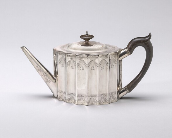 The spout of this silver teapot faces our left and the black handle our right in this photograph. The body of the teapot would be oval shaped if viewed from above. The sides of the teapot are straight and fluted to create vertical columns that curve inward. The shallowly domed lid has a flat, black, disk-like knob. The long, straight, narrow spout tapers from the base of the teapot to the opening, which is level with the top of the teapot. The handle to the right curves like the right half of a heart and flares upward where the thumb would rest at the top of the curve. The handle is affixed to the body of the teapot with short silver sleeves. The body is engraved along the top and bottom with decorative bands of garlands and vegetal scrolls. A rose-like flower and leaves are incised near the base on the side facing us.