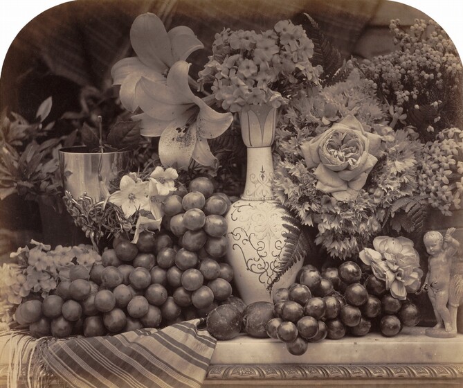 Grapes and flowers arranged on a tabletop in front of a dark background fill this horizontal photograph. The image is monochromatic like a black and white photograph but is printed in warm tones of golden and dark browns. The tabletop runs parallel and close to the bottom edge of the composition. It seems to be made of marble and a striped cloth folds over the front edge of the table to our left. Dark grapes are bunched around a vase at the middle of the composition. Its tall oval body and long, tapering neck are painted with decorative scrolls and curlicues. A few pieces of small round fruit, perhaps plums, rest on the table in front of the vase. A silver goblet to the left and a small figurine of a young boy with a sheaf of wheat to our right are almost lost in the profusion of roses, lilies, hyacinth, and other flowers that fill the space around and behind the objects. The top corners of the photograph are curved to create an arched effect for the composition.