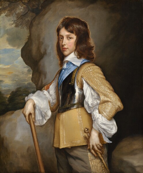 Shown from the hips up, a boy with pale, peachy skin and copper-brown hair stands in front of a rocky outcropping with a narrow view into a distant landscape to our left in this vertical portrait painting. His body faces our left almost in profile, and he turns his face to look at us with dark brown eyes under faint brows. He has a long, straight nose, a pointed chin, flushed cheeks, and his full, rose-pink lips are slightly parted. His dark hair falls in waves to the wide, flat, white collar at his neck. He wears a golden yellow doublet brocaded with shimmering threads, possibly silver and gold. The sleeves split along the inner arm to reveal a white shirt with voluminous, puffy sleeves that end with wide, transparent cuffs like floppy saucers around each wrist. Over the jacket, he wears a gleaming, pewter-colored breastplate with a topaz-blue sash crossing his chest. His peanut-brown pants are also sewn or woven with metallic thread in stripes down the side of the leg. The boy rests his right hand, farther from us, on a staff or walking stick propped in front of him. His other hand rests around the gold hilt of a rapier, a thin sword at his left hip, closer to us. Behind the boy, the view is almost entirely taken up with a slate-gray boulder, or it could be the entrance of a cave. A view onto the landscape along the left edge of the composition has a few treetops beneath pale yellow clouds against an ice-blue sky.