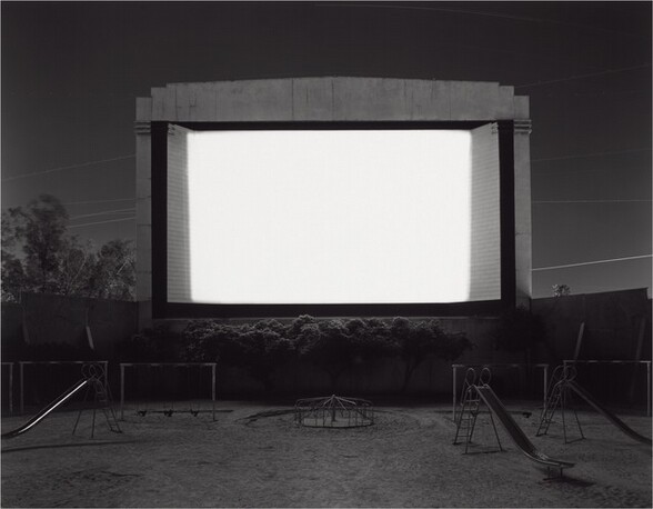A movie screen is bright white against a dark gray sky, trees, and a playground in this horizontal, black and white photograph. The screen takes up about a quarter of the overall composition, just above the center of the picture. It is raised off the ground, perhaps on a wall or stage that we cannot see. Light from the screen brushes the tops of trees or shrubs growing near the bottom edge. Three sliding boards, several swing sets, and a merry-go-round fade in and out of shadow around the screen’s glow. Power lines crisscross the dark, clear sky behind the screen.